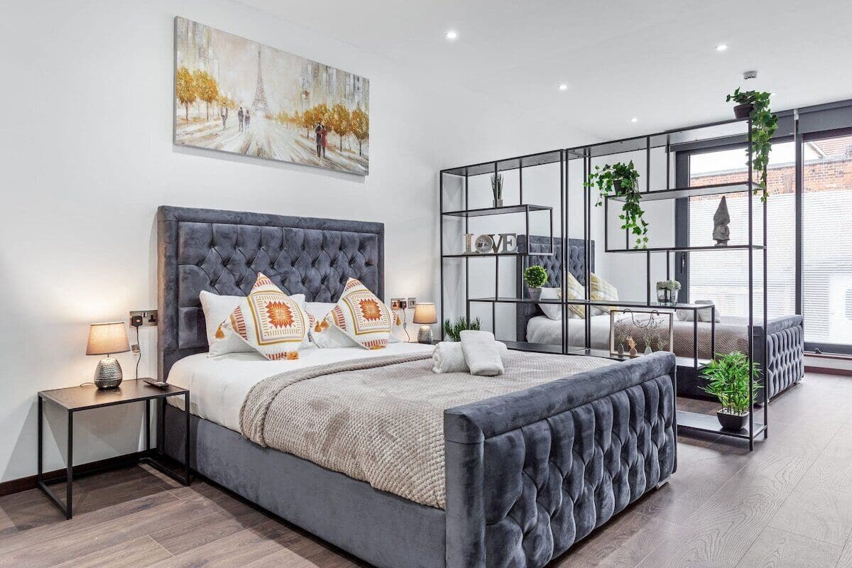 A modern bedroom with a large bed, bedside tables, a decorative mirror, and a cityscape painting.