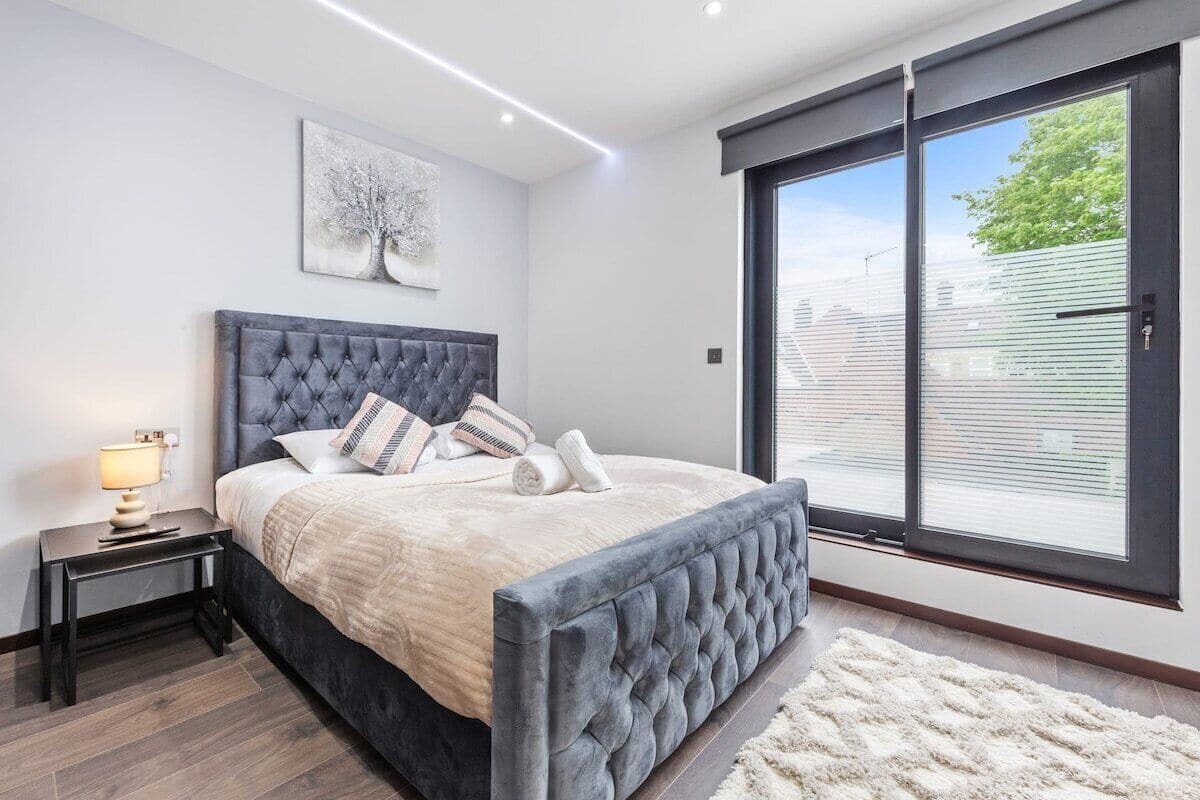 A modern bedroom with a large bed, a nightstand, a lamp, a window with blinds, and a painting on the wall.
