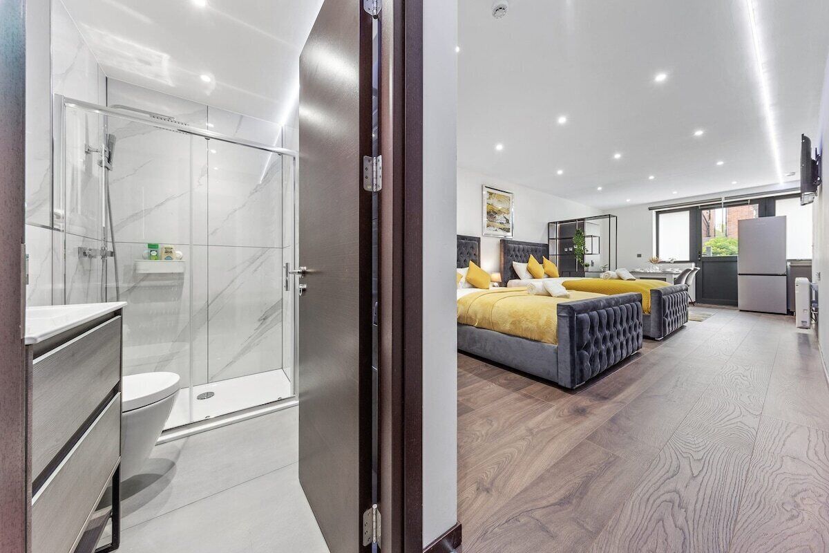 Una habitación de hotel moderna con cabina de ducha de vidrio, inodoro independiente, cama con cabecero y un amplio ventanal que proporciona luz natural.