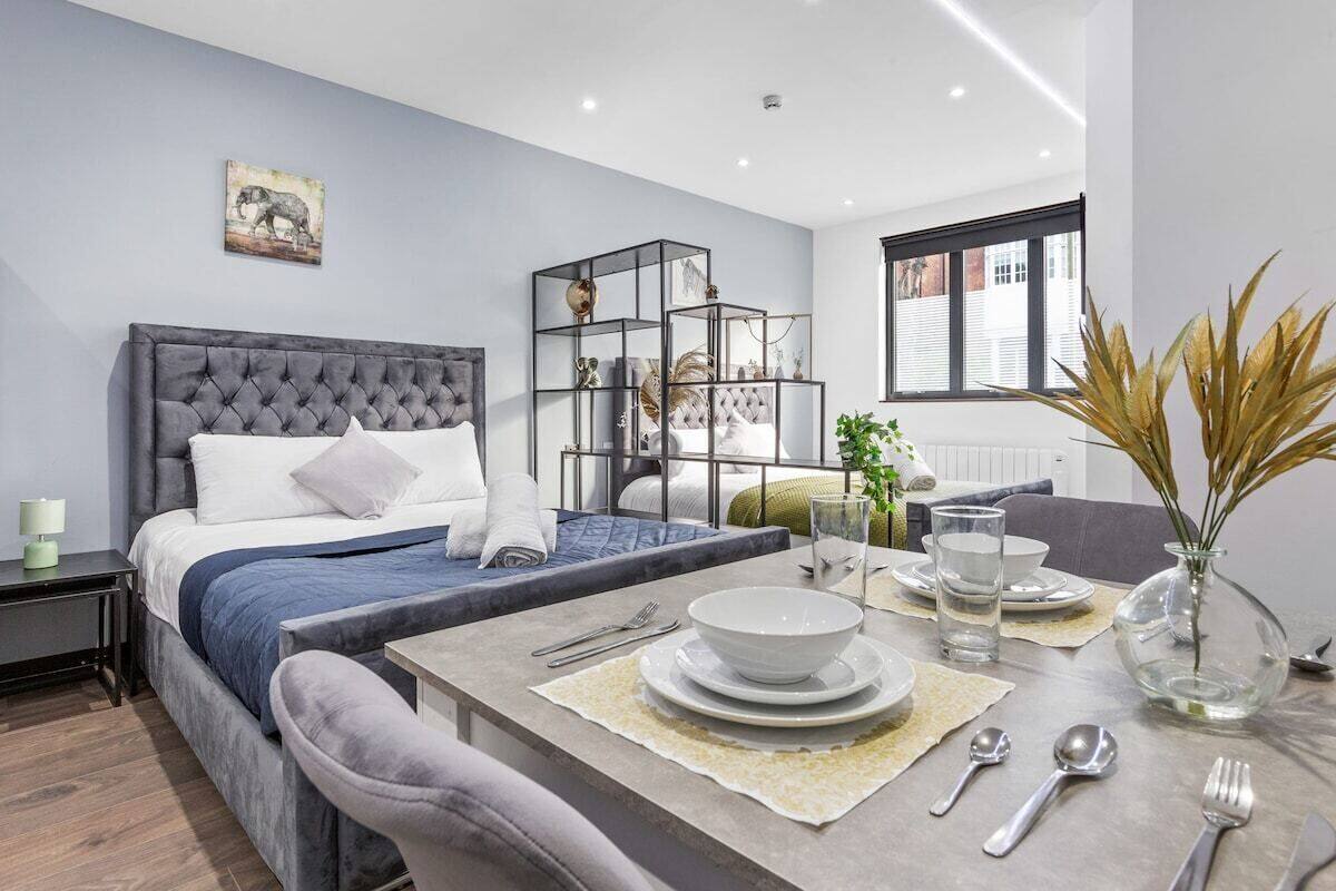 A modern, well-lit bedroom with a tufted headboard bed, a dining area with a table set for two, and a large window providing natural light.