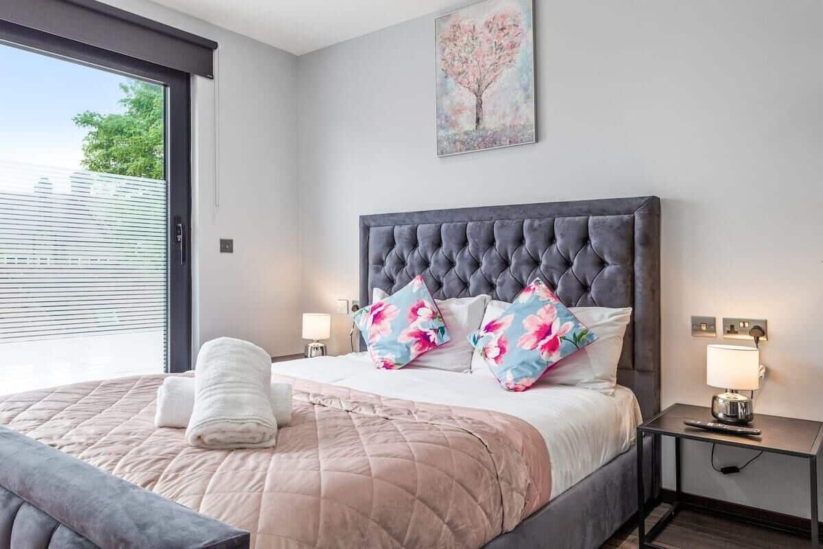 A bedroom with a large bed, a grey upholstered headboard, and a window with blinds.