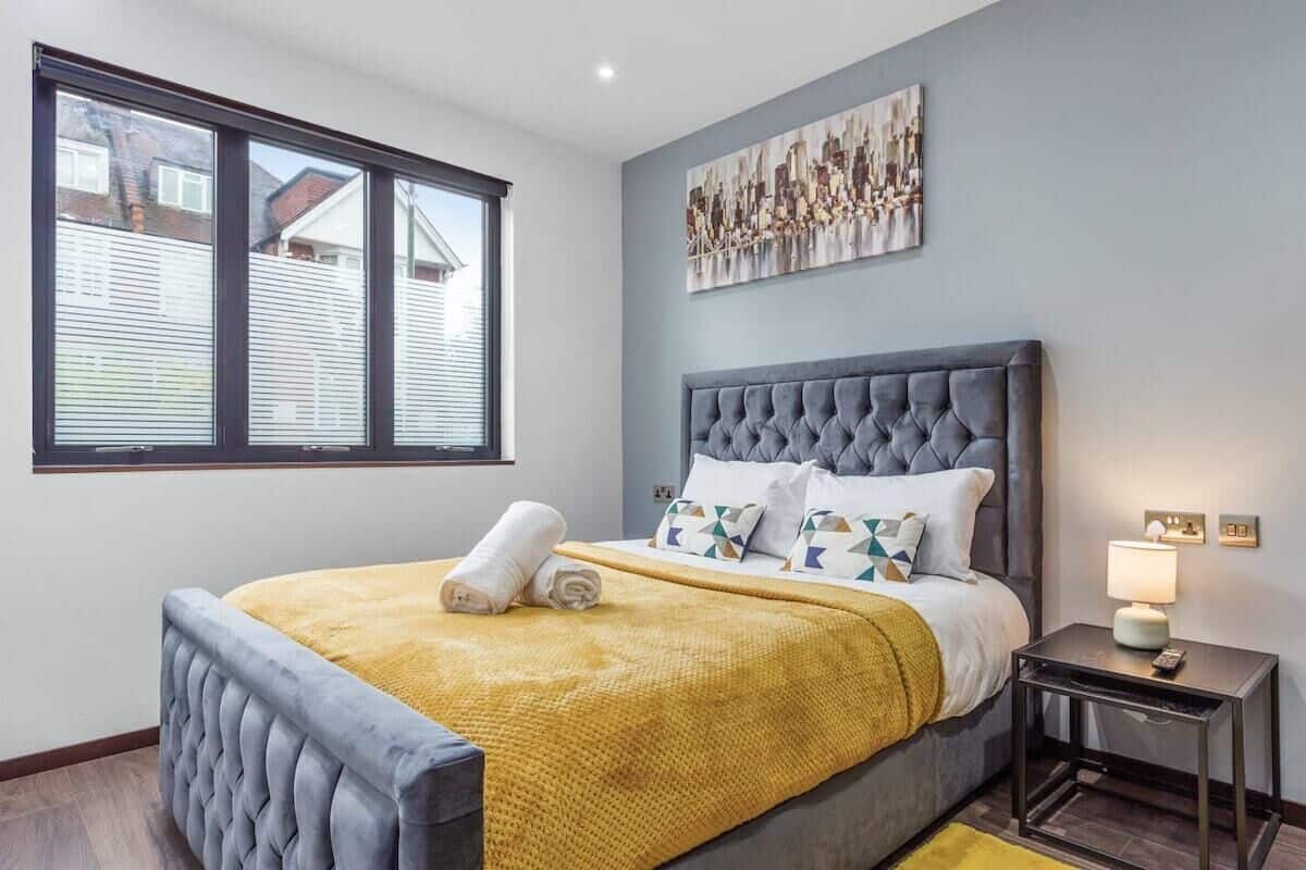A bedroom with a large bed, a window, a cityscape painting, and a nightstand with a lamp.
