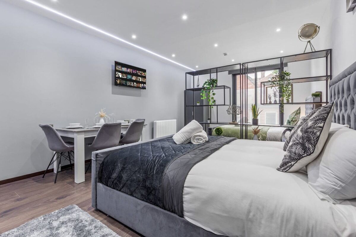 A modern bedroom with a large bed, a desk, and a chair. There is a bookshelf with books and a plant.