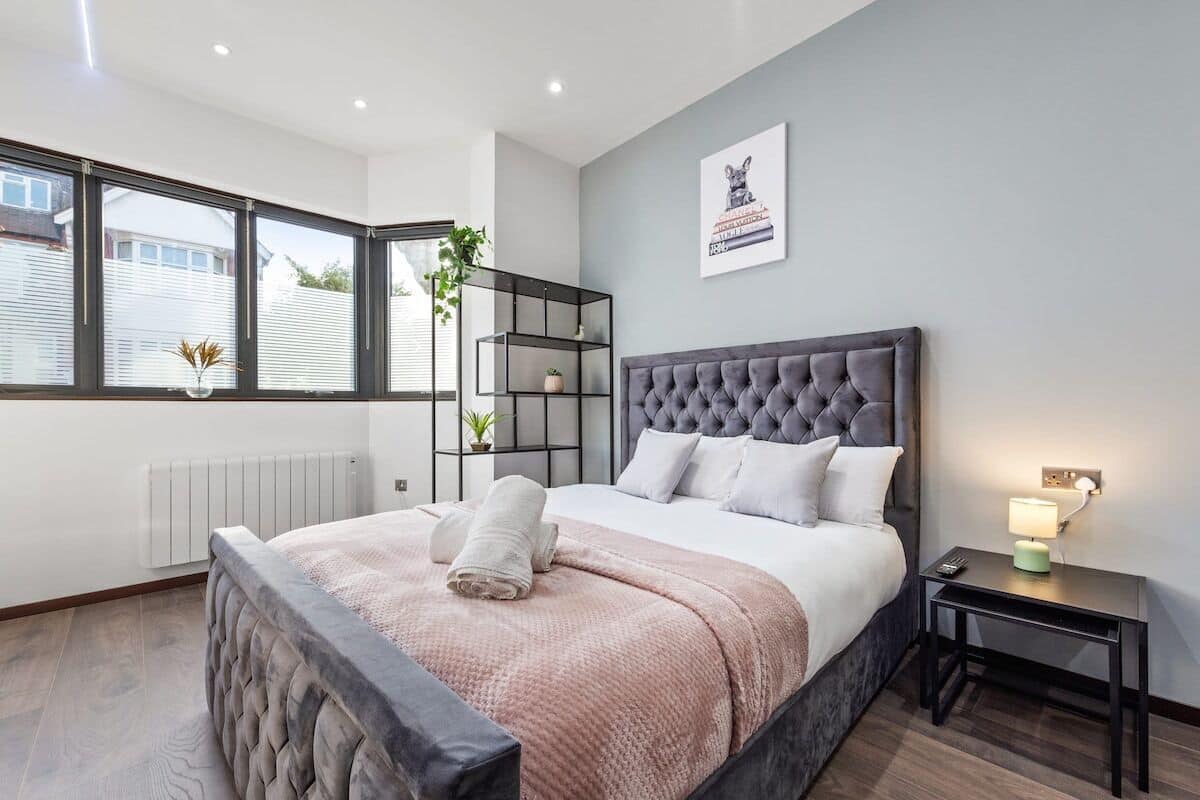 A modern bedroom with a large bed, a bedside table with a lamp, a window with blinds, and a wall-mounted shelf with plants.