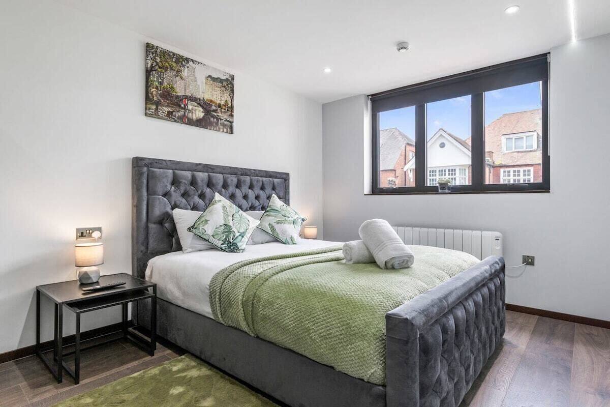A bedroom with a large bed, a nightstand, a window with a view of houses, and a painting on the wall.