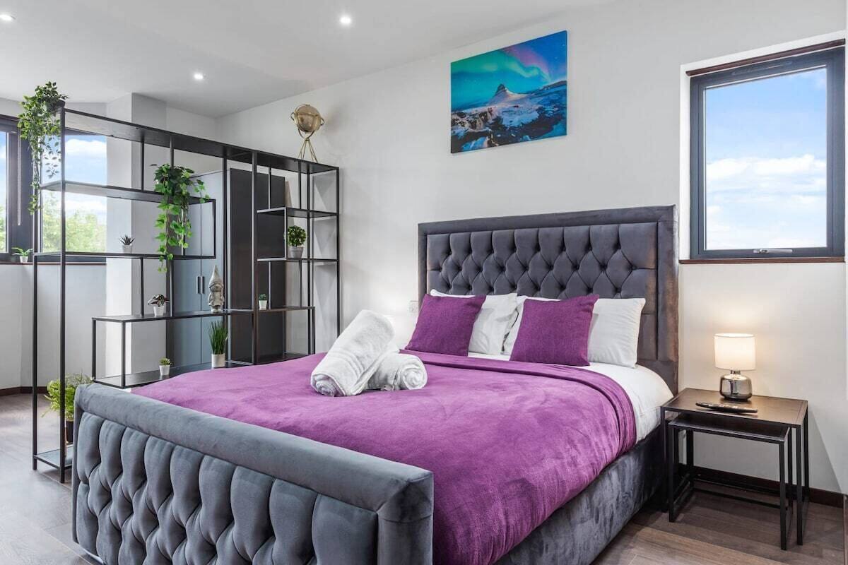 A modern bedroom with a tufted headboard, a purple bedspread, and a view of the sky through a window.