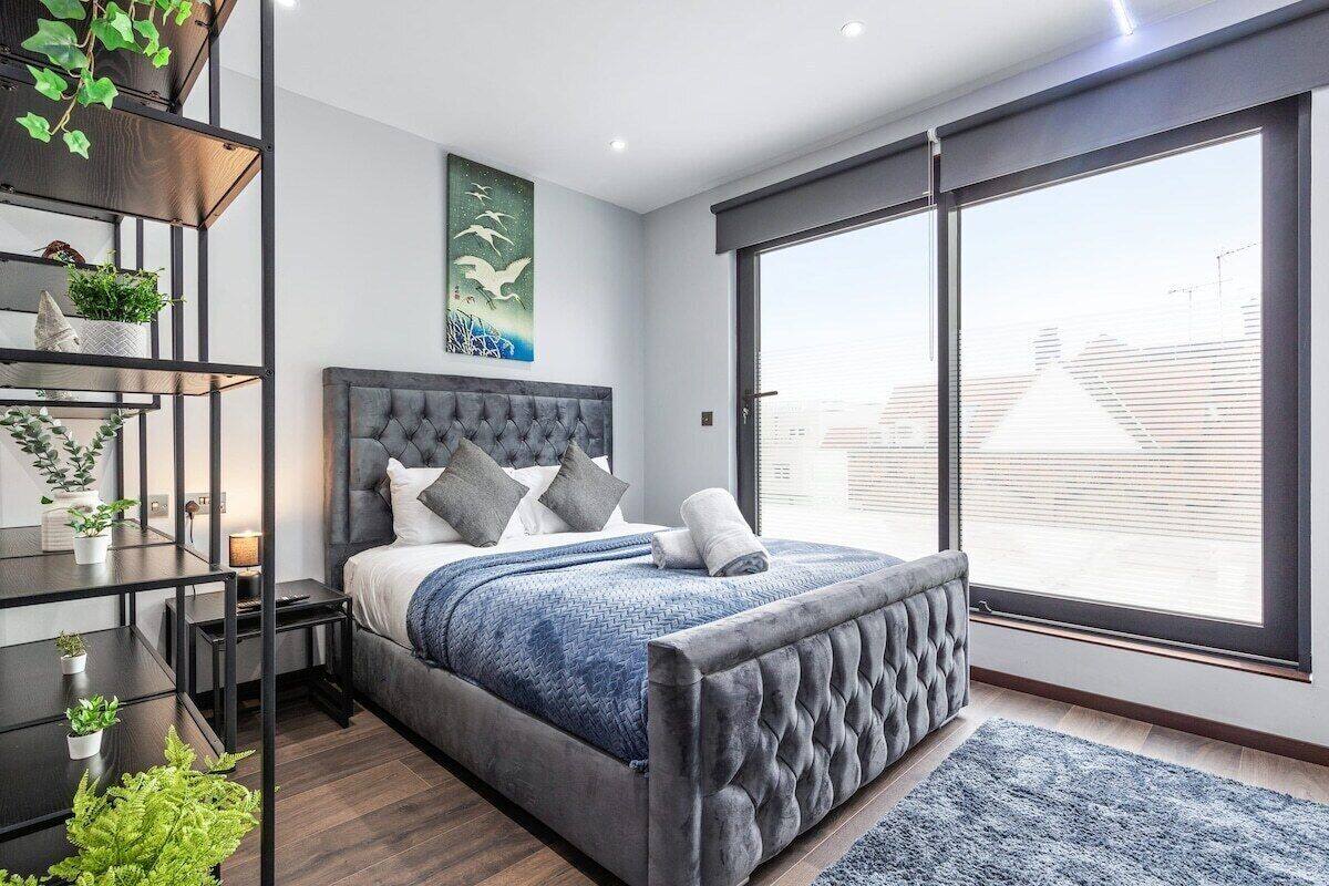 A modern bedroom with a large bed, a grey upholstered headboard, a wall-mounted shelf with plants, and a large window offering a city view.