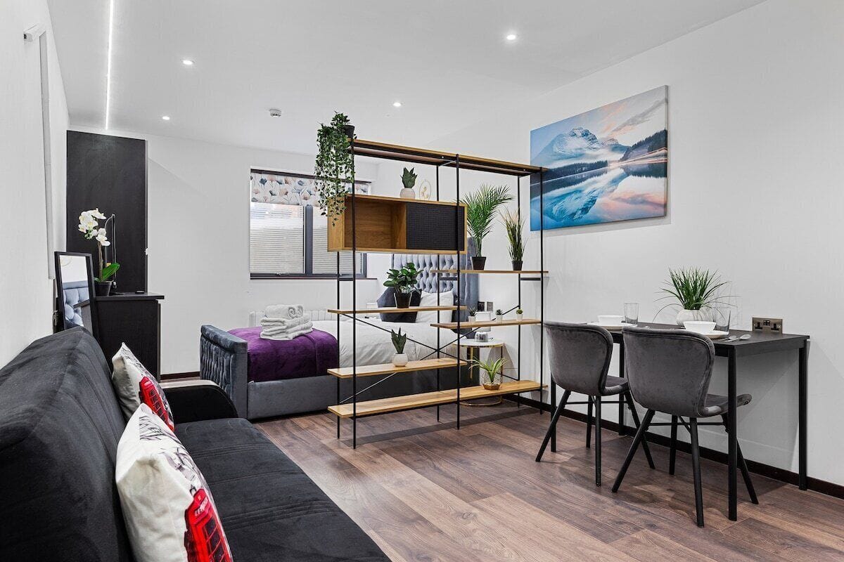 A modern living room with a sofa, a desk with two chairs, a shelving unit with plants, and a large painting on the wall.