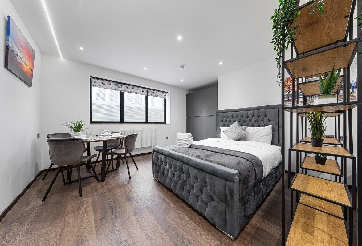 A modern bedroom with a large bed, a desk, and a chair. There is a window with blinds, a radiator, and a plant on a shelf.