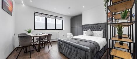 A modern bedroom with a large bed, a desk, and a chair. There is a window with blinds, a radiator, and a plant on a shelf.