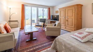 A well-lit room with a bed, a desk, a chair, a wardrobe, and a view through the glass doors.
