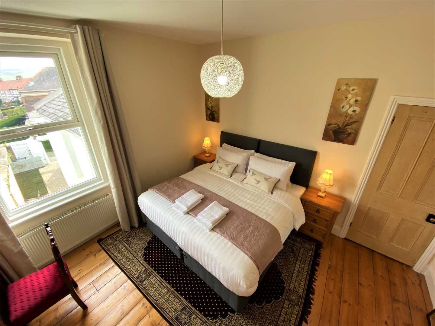 A neatly made bed with pillows and a blanket, a hanging light fixture, a painting on the wall, and a door.