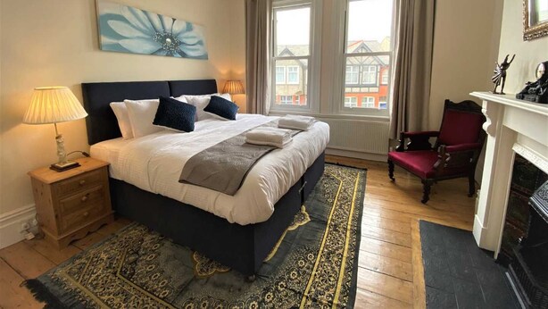 A bedroom with a bed, bedside lamps, a rug, a chair, a fireplace, and a view of buildings through the window. - Tre@Westminster_Residence - Spacious Period Townhouse in Douglas with EV Charger (Douglas)