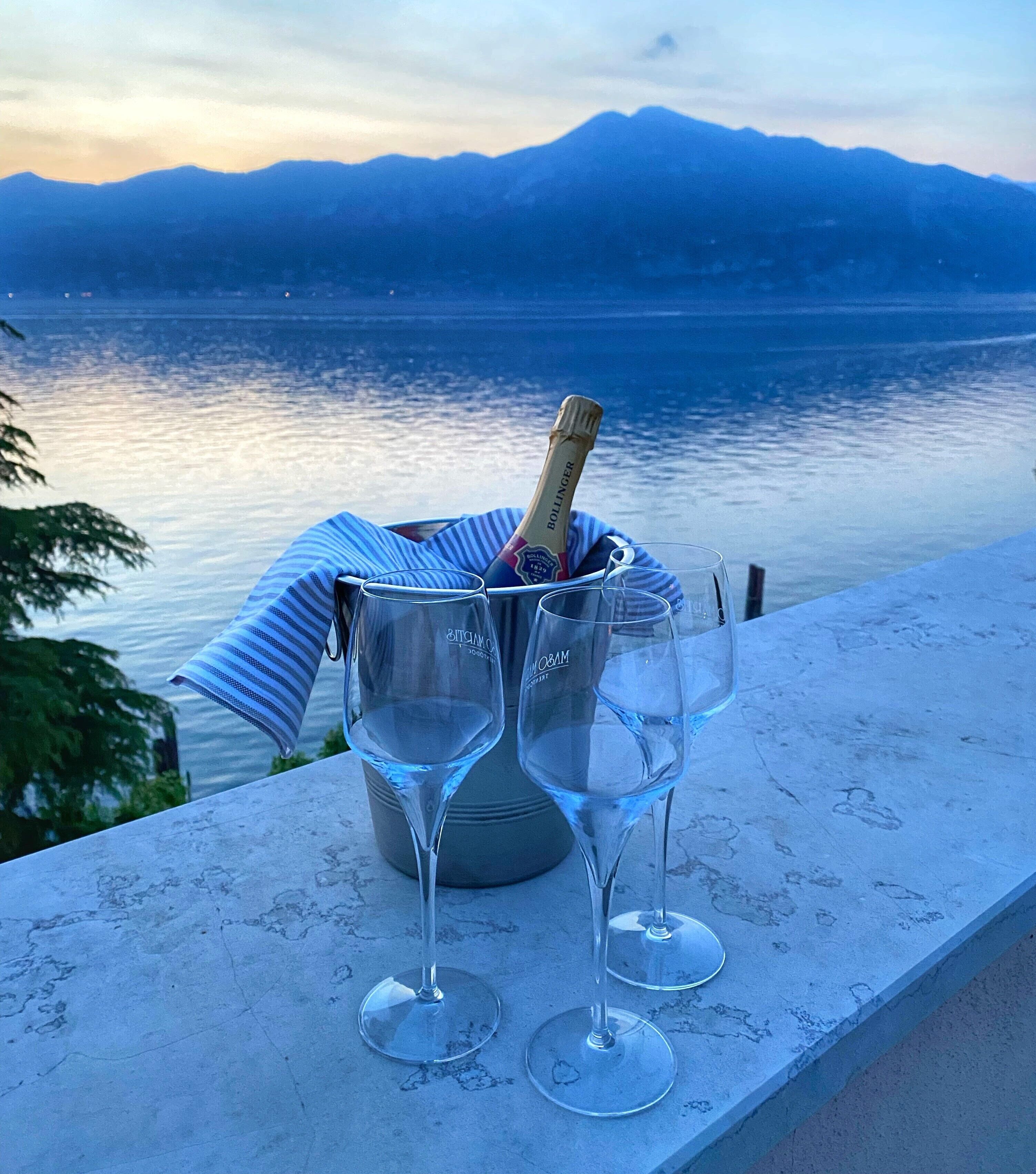 A champagne bottle and two glasses on a balcony overlooking a lake with mountains in the background.