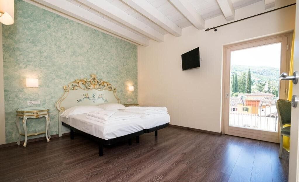 A bedroom with a bed, a nightstand, a wall-mounted TV, and a door leading to a balcony with a view of buildings and trees.