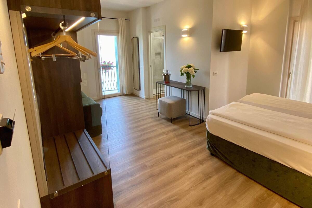 Una habitación de hotel con un banco de madera, una cama, una mesita con un florero y un televisor montado en la pared.