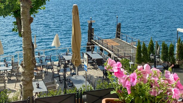 A seaside restaurant with outdoor seating overlooking a lake, surrounded by mountains. - Hotel Pai (Torri del Benaco)