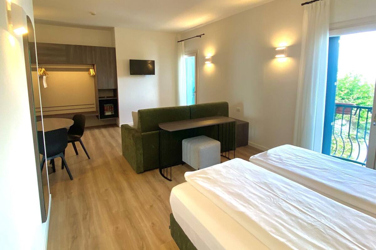 Una habitación de hotel moderna con una cama grande, un sofá verde, una mesita pequeña y un escritorio. Hay un televisor montado en la pared, y se ve un balcón con barandilla a través de la ventana.
