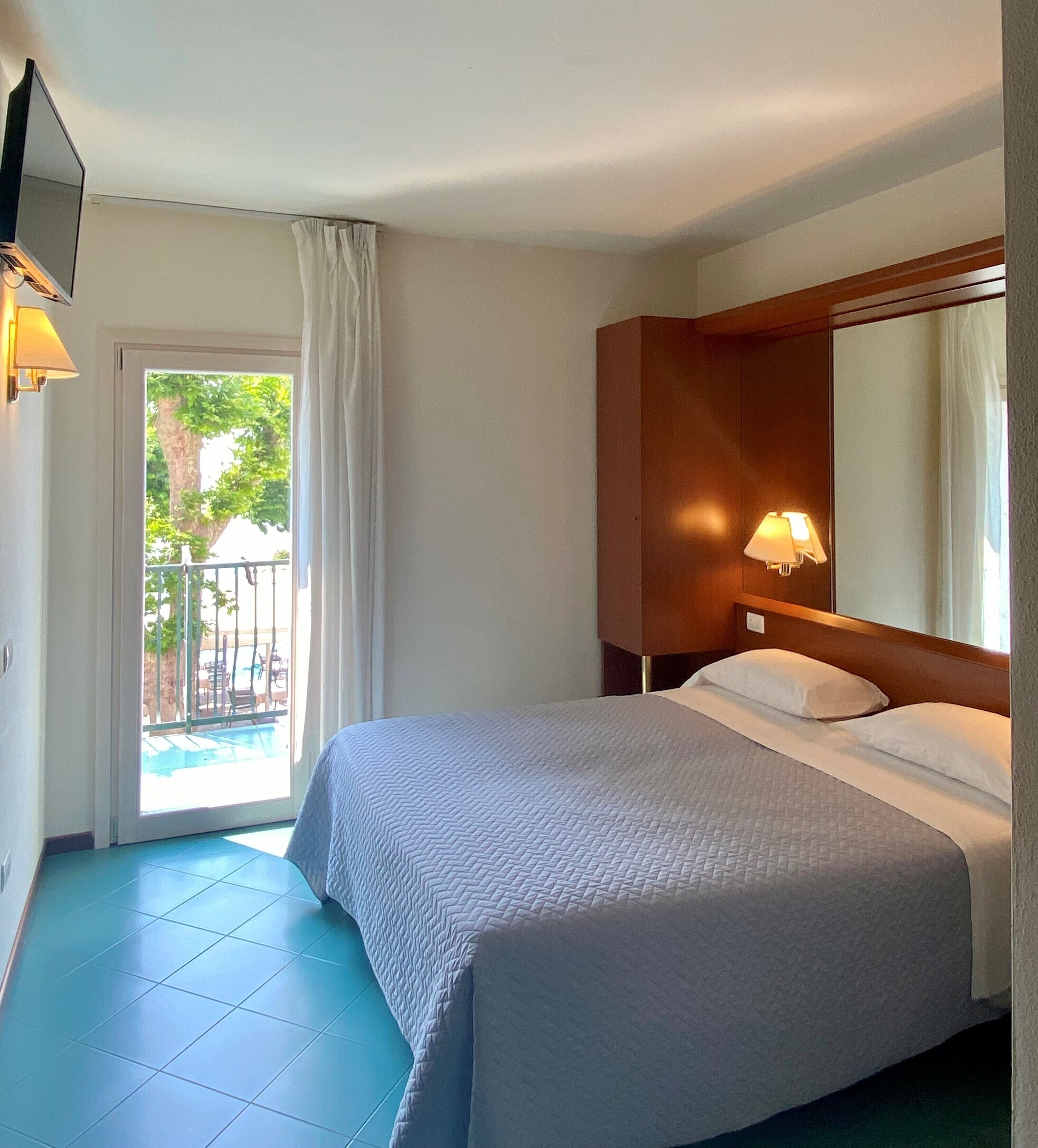 Une chambre d’hôtel comprenant un lit, une table de chevet avec une lampe, un grand miroir et une porte-fenêtre coulissante donnant sur un espace extérieur.