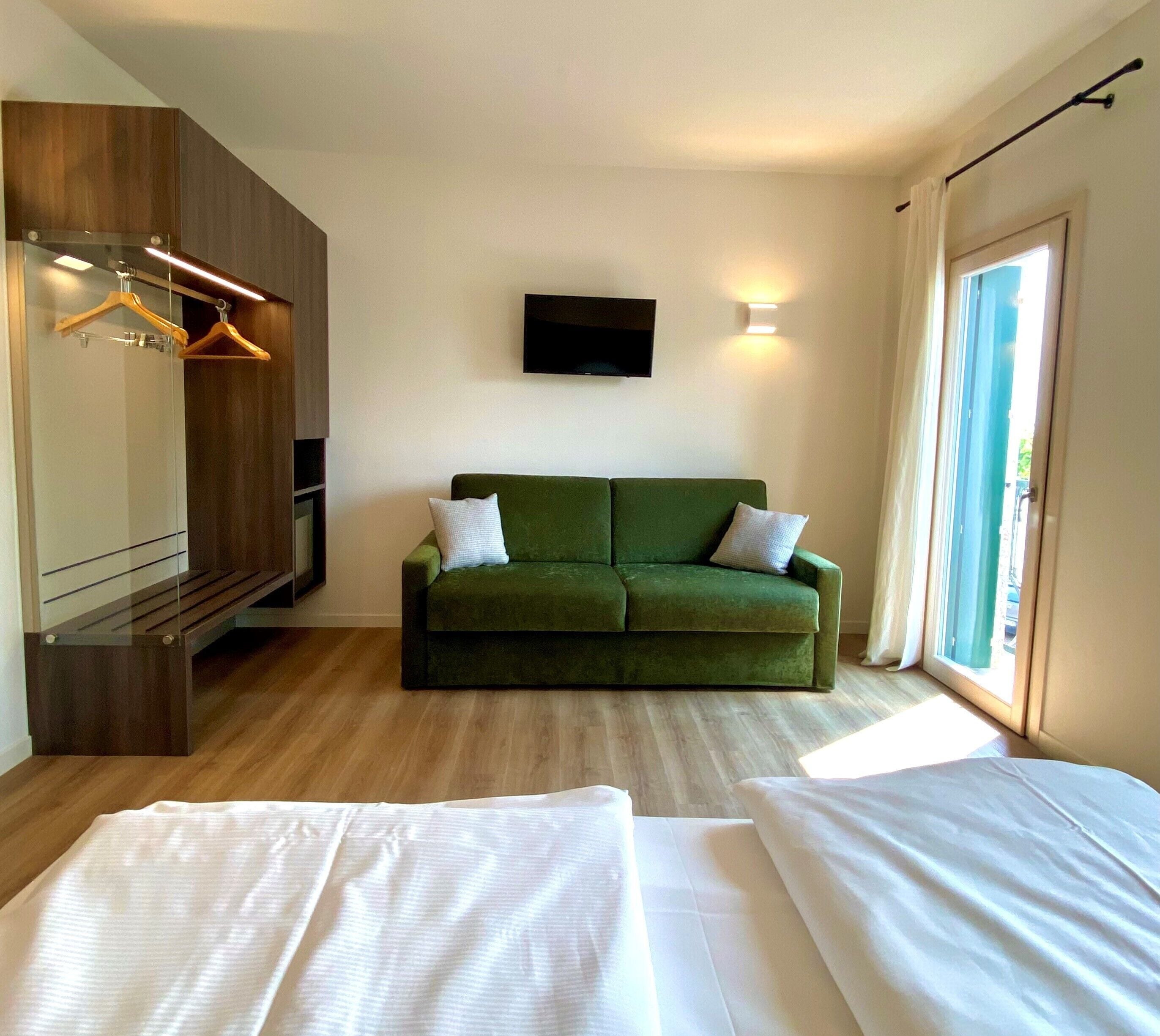 Une pièce avec un canapé vert, un téléviseur à écran plat fixé au mur, une armoire en bois et une grande fenêtre avec des rideaux.