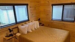 Een houten kamer met een bed, twee ramen met zonwering, een nachtkastje en een lamp.