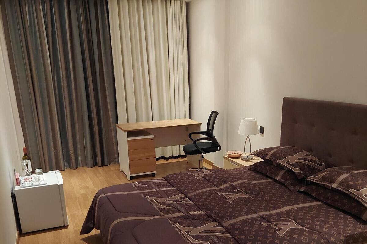 A bedroom with a large bed, a desk, a chair, a nightstand, and a lamp.