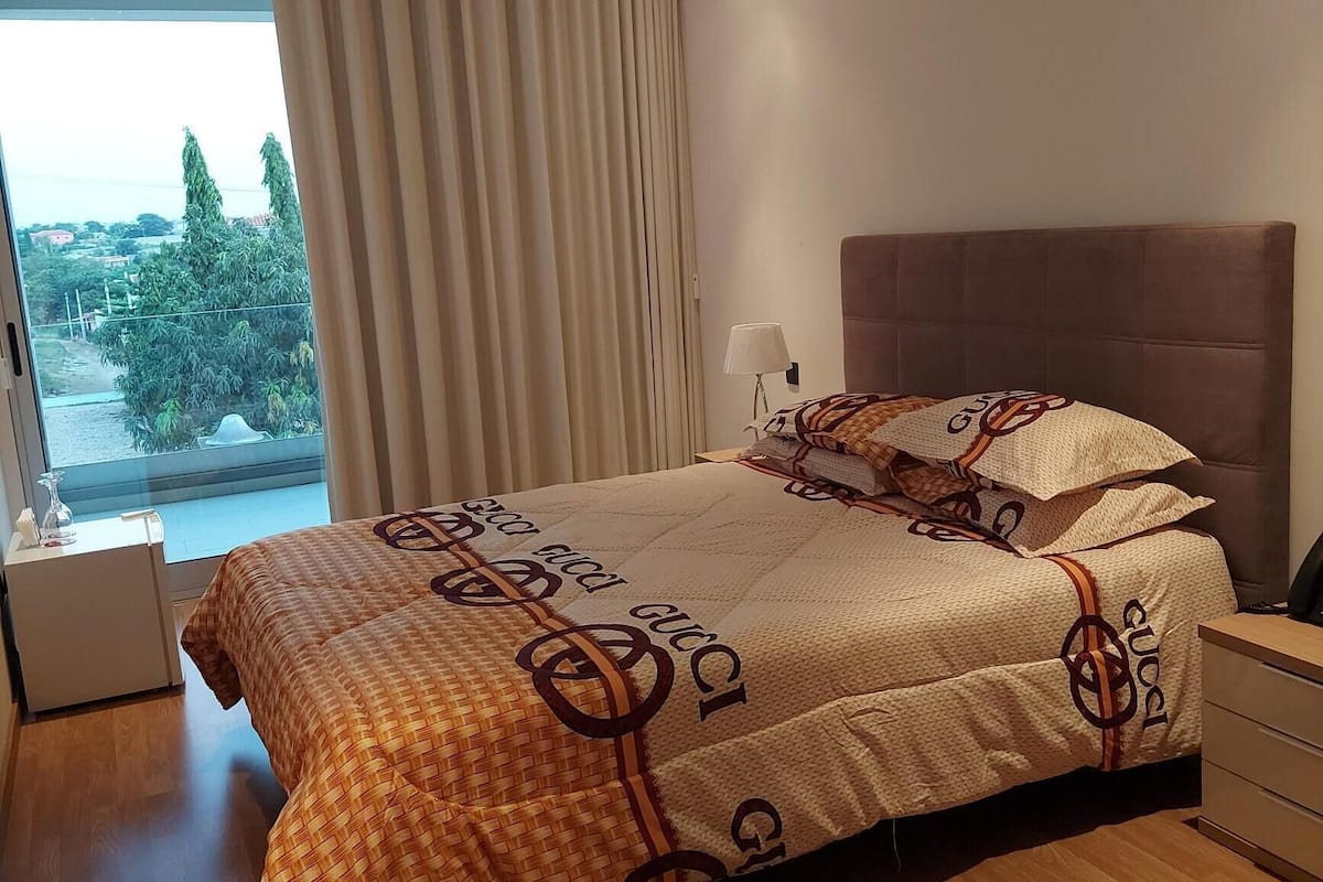 A modern bedroom with a large bed, a nightstand, a lamp, and a view of the outdoors.