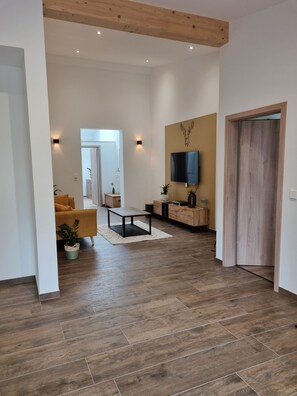 Een ruime woonkamer met houten vloer, een bank, salontafel en een aan de muur bevestigde televisie.