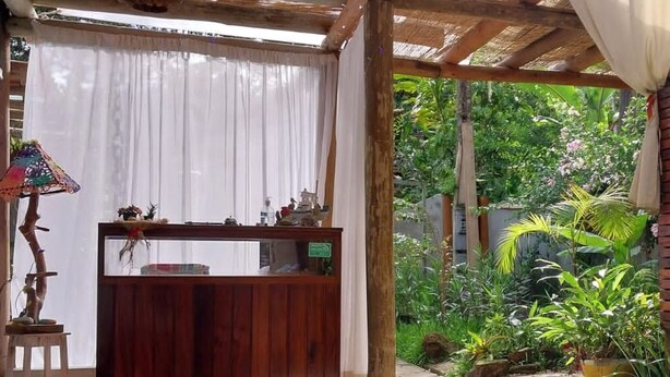 Un banc en bois placé sous une pergola, avec un rideau blanc en arrière-plan, entouré d’une végétation luxuriante et de plantes en pot.