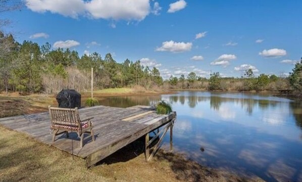 A wooden dock extending over a calm lake, with a chair and grill on the dock.