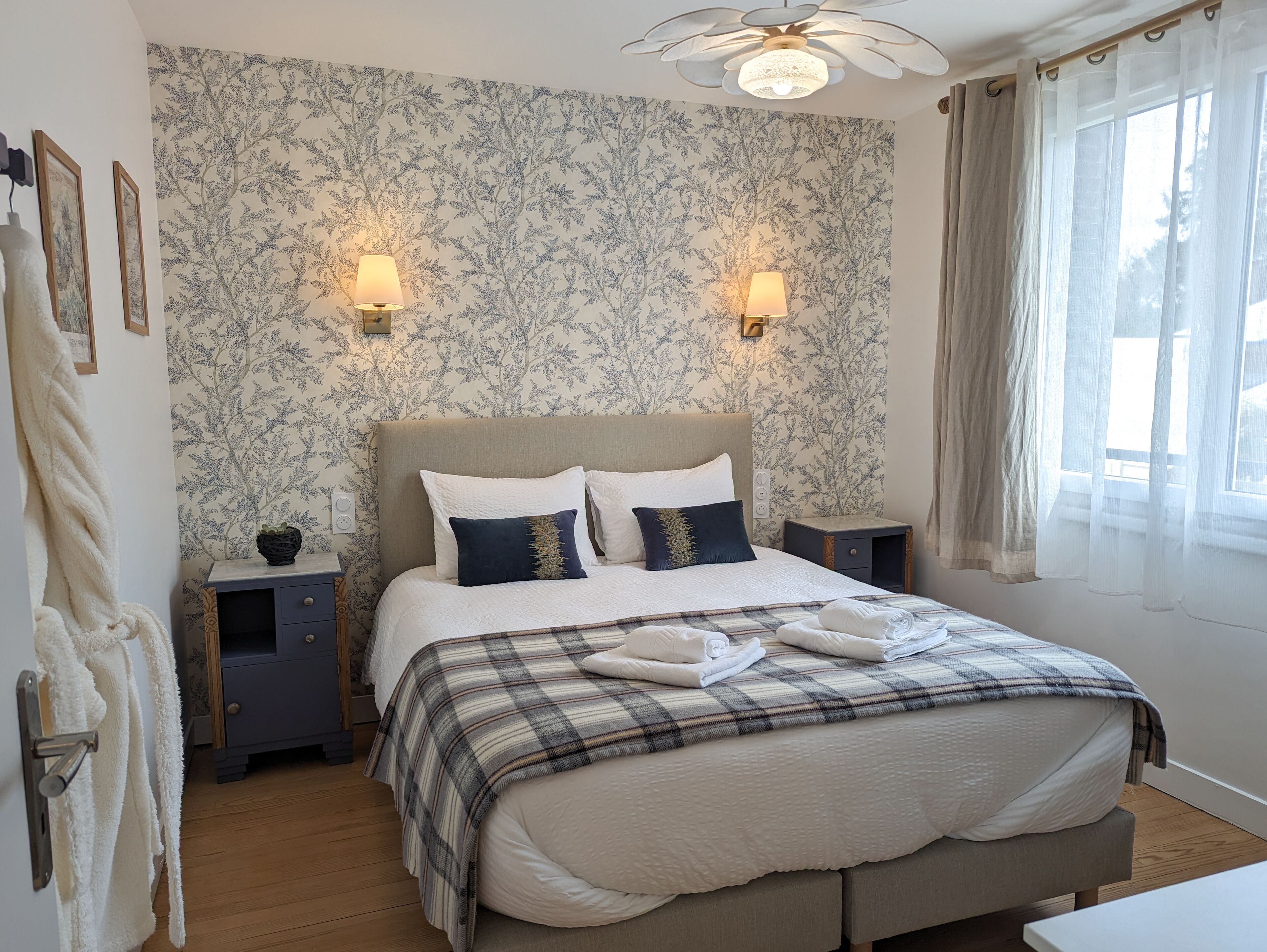 Ein ordentlich gemachtes Bett mit einer Karo-Decke, zwei Nachttischlampen, einem Nachttisch und einem Fenster mit durchsichtigen Vorhängen.