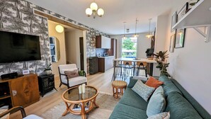 A modern living room with a large flat-screen TV, a green sofa, a coffee table, and a dining area with a kitchen in the background. - Le Cémarose Charming cottage in Champagne near Epernay (Épernay)
