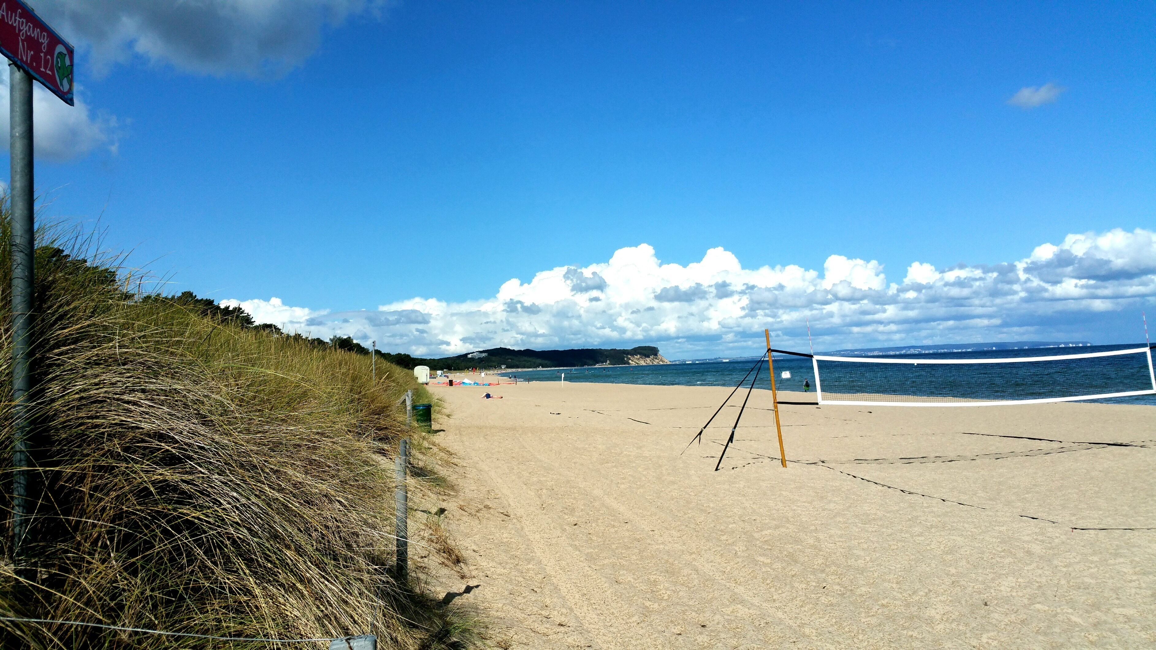 A beach volleyball court with a net, a sandy beach, and a grassy area with a sign.