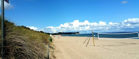 A beach volleyball court with a net, a sandy beach, and a grassy area with a sign.