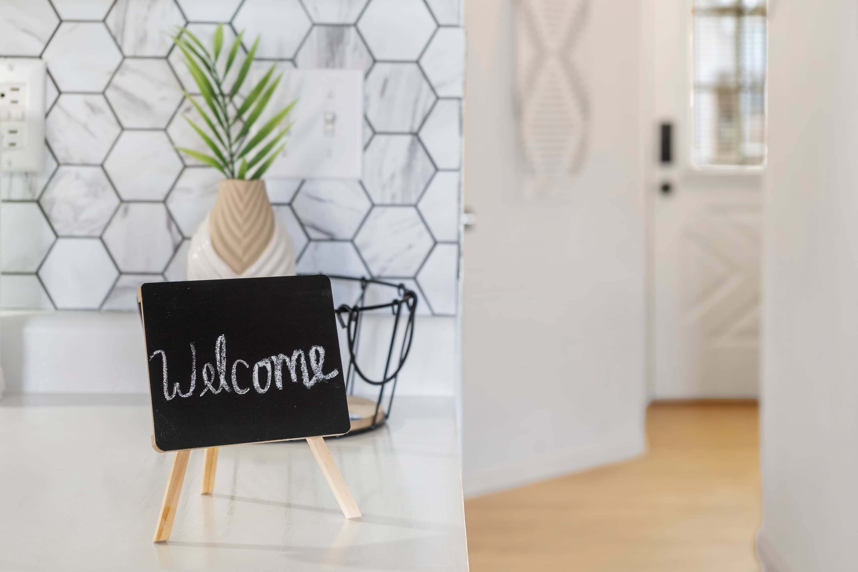 Eine Tafel mit dem Schriftzug „Welcome“, auf einem Ständer in einer modernen Küche mit sechseckigen Fliesen und einem Blumentopf.