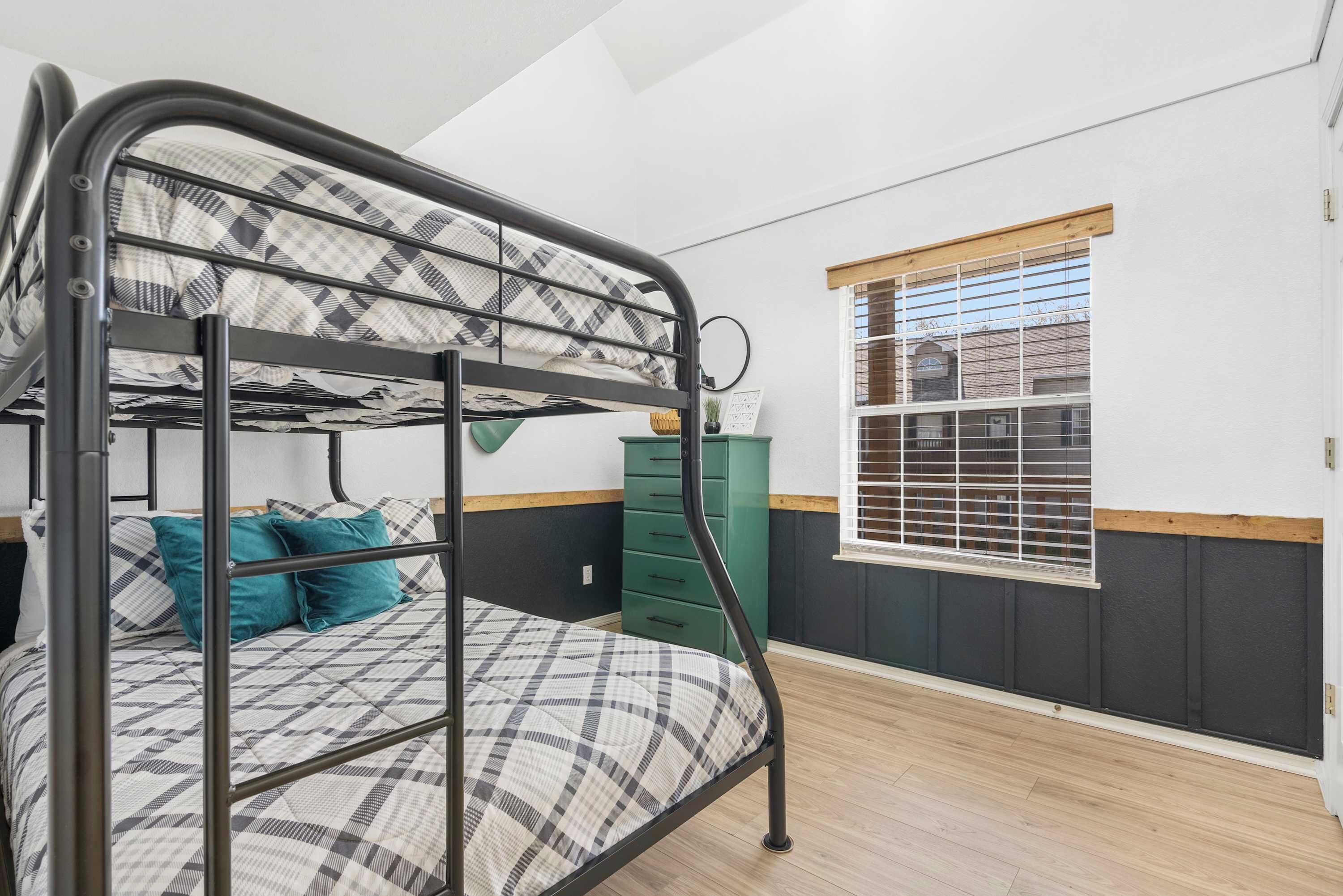 A bunk bed with a checkered bedspread and teal pillows, a green dresser, and a window with blinds.