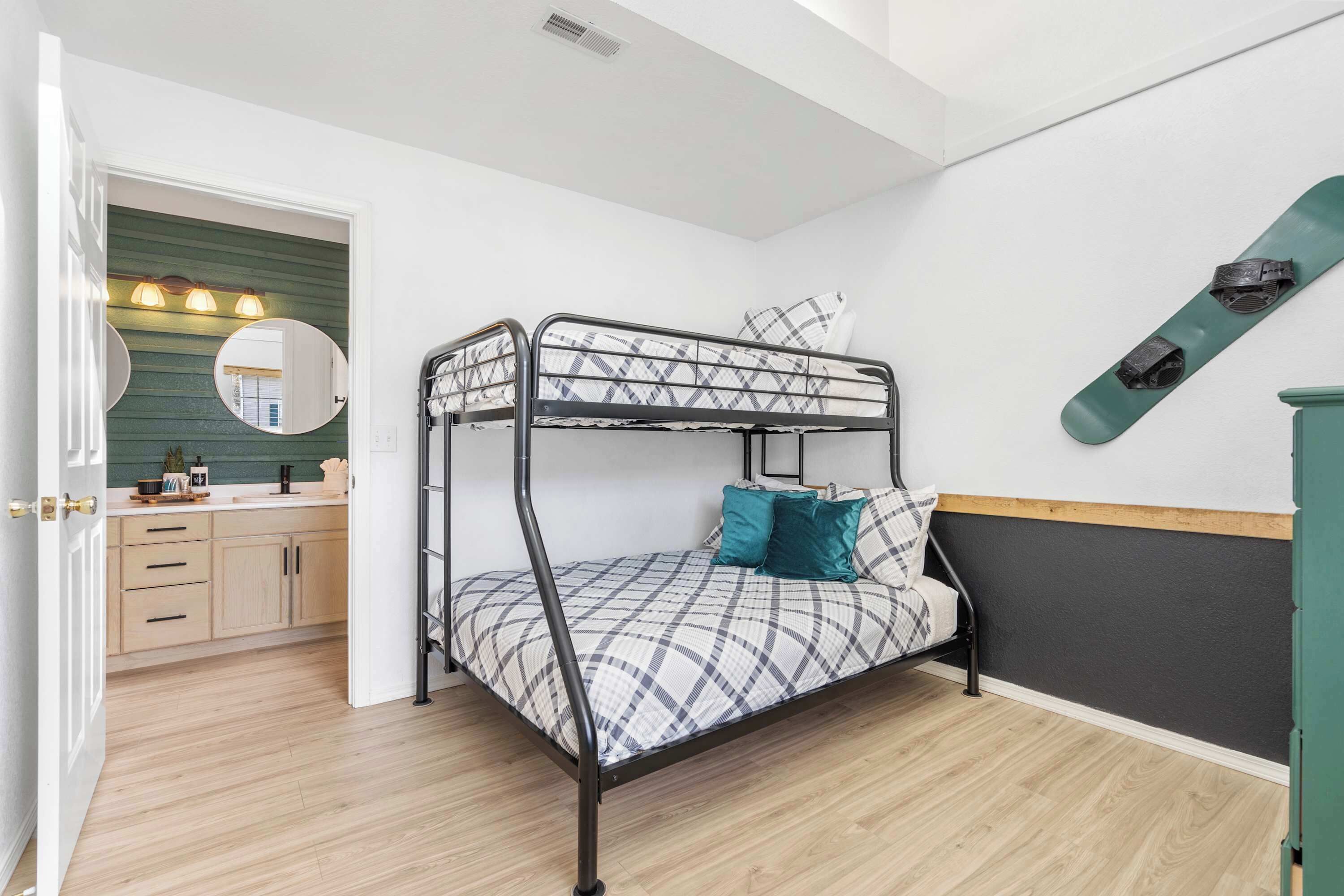 A bunk bed with a checkered bedspread and teal pillows, a green snowboard leaning against the wall, and a kitchen area with wooden cabinets and a green backsplash.