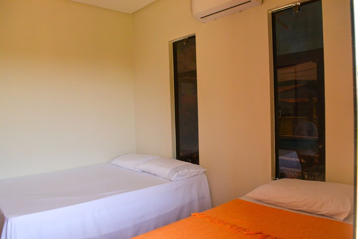 A room with two beds, one with a white bedspread and the other with an orange bedspread. There is a large window on the right side of the room.