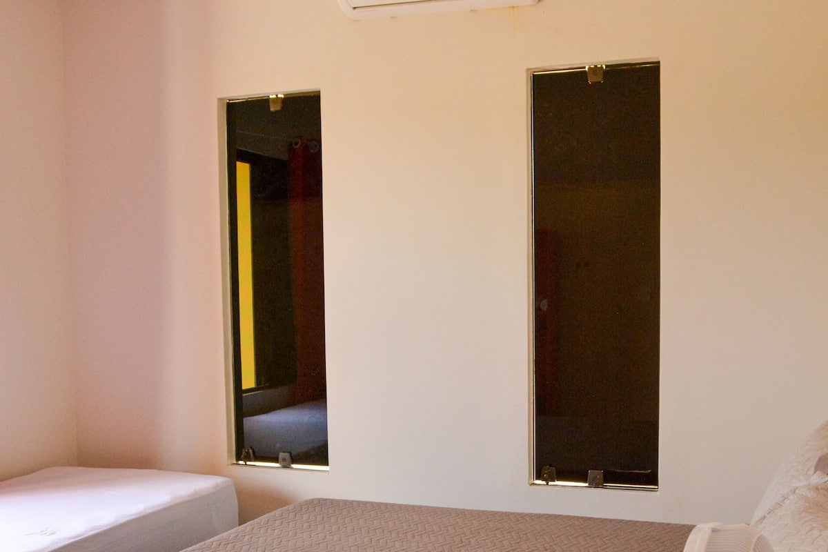 A bedroom with a bed, a standing fan, an air conditioner, and two windows.