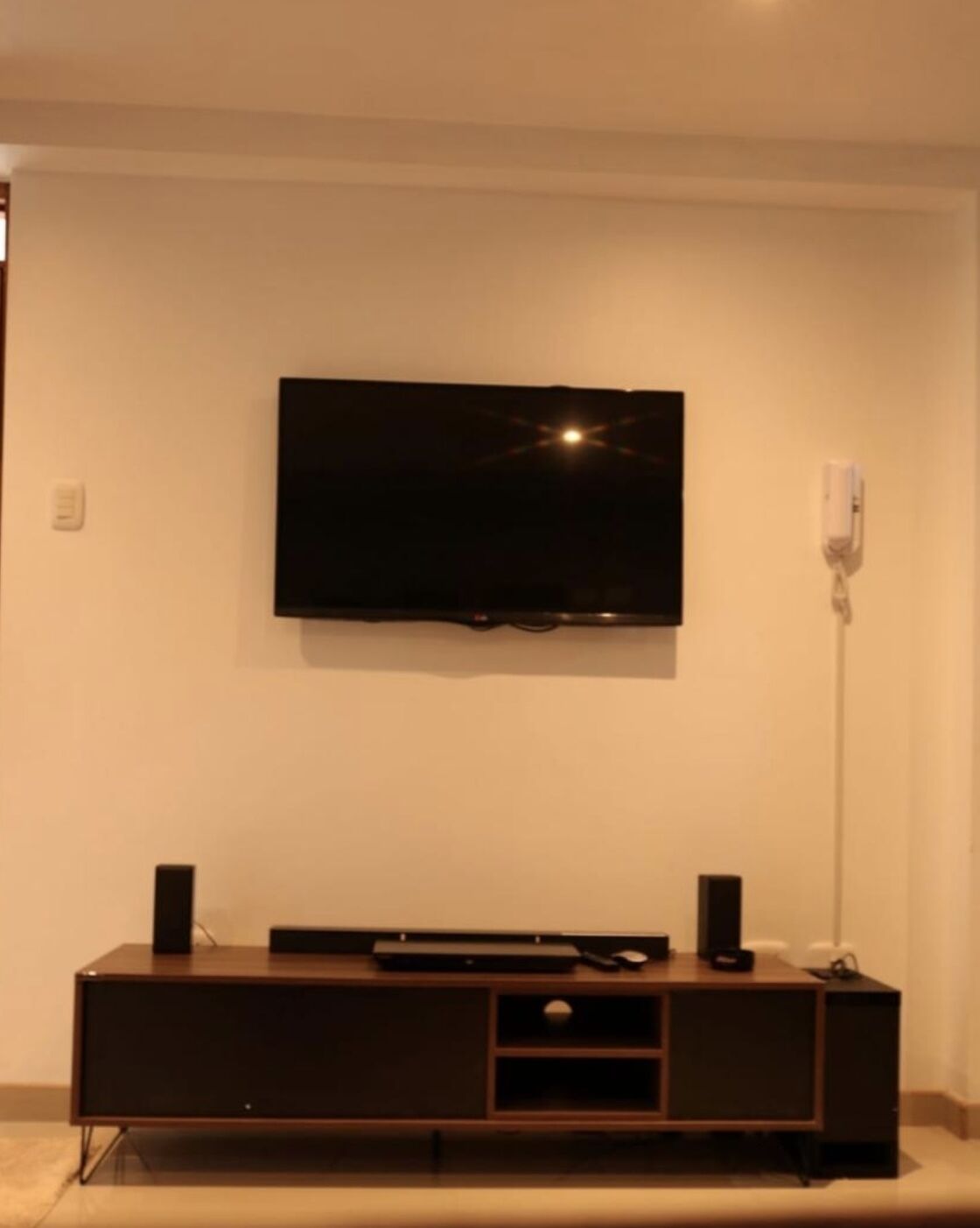 A flat-screen TV mounted on a wall above a dark wooden entertainment center with a remote control and a speaker on each side.