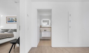 A modern hotel room with a bed, a desk, a bathroom with a mirror and sink, and a door leading to another room.