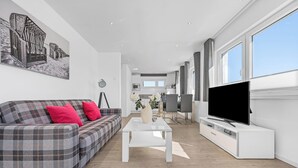A modern living room with a sofa, coffee table, TV, and a kitchen in the background. - Strandkrabbe - 1st row on the water & beach, ground floor, shower room, terrace (Olpenitz)
