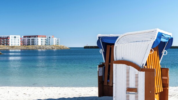 A beach chair with a blue and white cover, a sandy beach, and a row of colorful buildings across the water. - Strandkrabbe - 1st row on the water & beach, ground floor, shower room, terrace (Olpenitz)