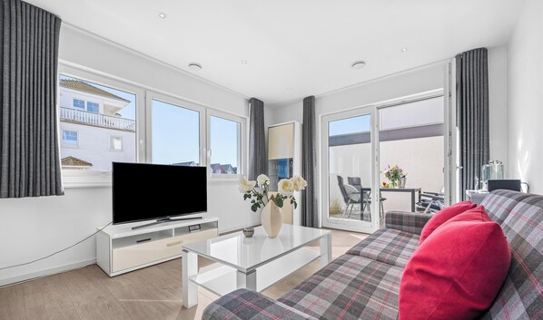 A modern living room with a flat-screen TV, a glass coffee table, a sofa with a plaid cover, and a balcony with a dining area.