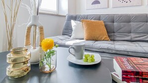 Un salon moderne avec un canapé gris, un vase blanc contenant des fleurs jaunes, un vase en verre avec des plantes vertes, une tasse grise et une assiette de raisins.