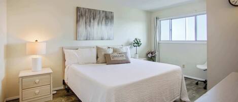 A neatly arranged bedroom with a large bed, a bedside table with a lamp, and a window with curtains.