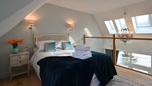 Ein Schlafzimmer mit schrägverdeckter Decke, einem Bett mit dunkelblauer Bettdecke, zwei Wandlampen, einer Pendelleuchte und Blick durch ein Fenster.