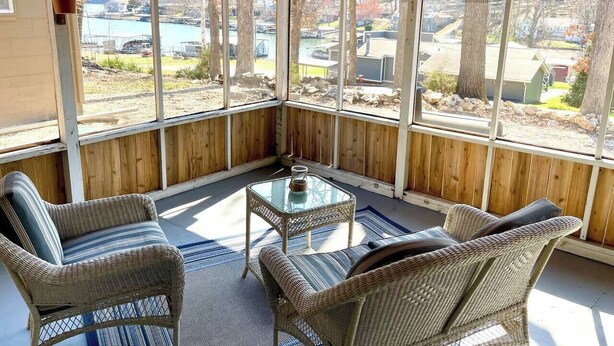 Eine überdachte Terrasse mit Korbmöbeln und einem Glastisch, mit Blick auf einen See und eine Wohngegend.