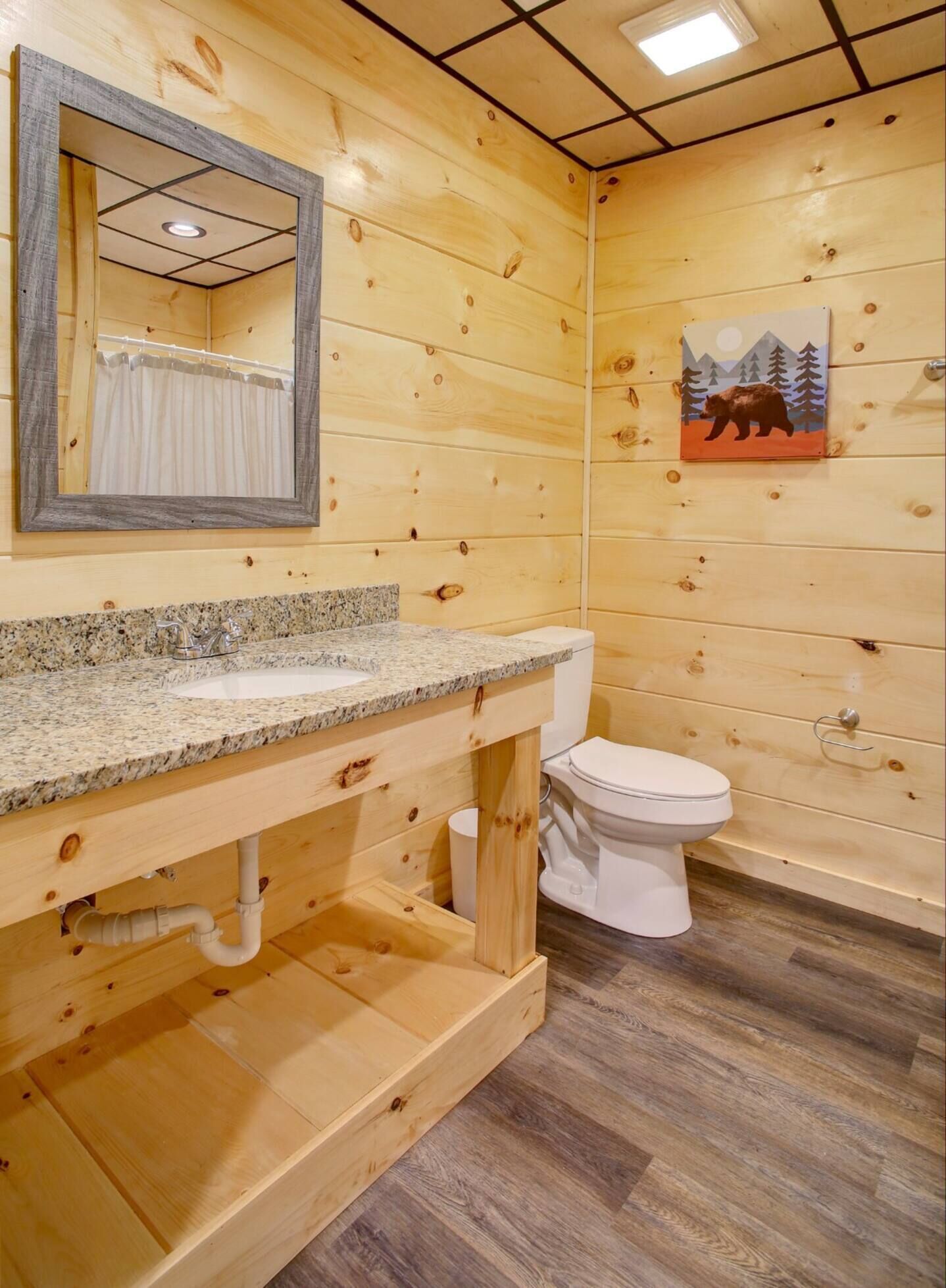 Baño de cabaña de madera con lavamanos de granito, inodoro blanco y espejo sobre el lavamanos.