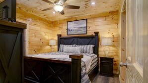 Un dormitorio de cabaña de madera con una cama, dos mesitas de noche, un ventilador de techo y un cuadro enmarcado en la pared.